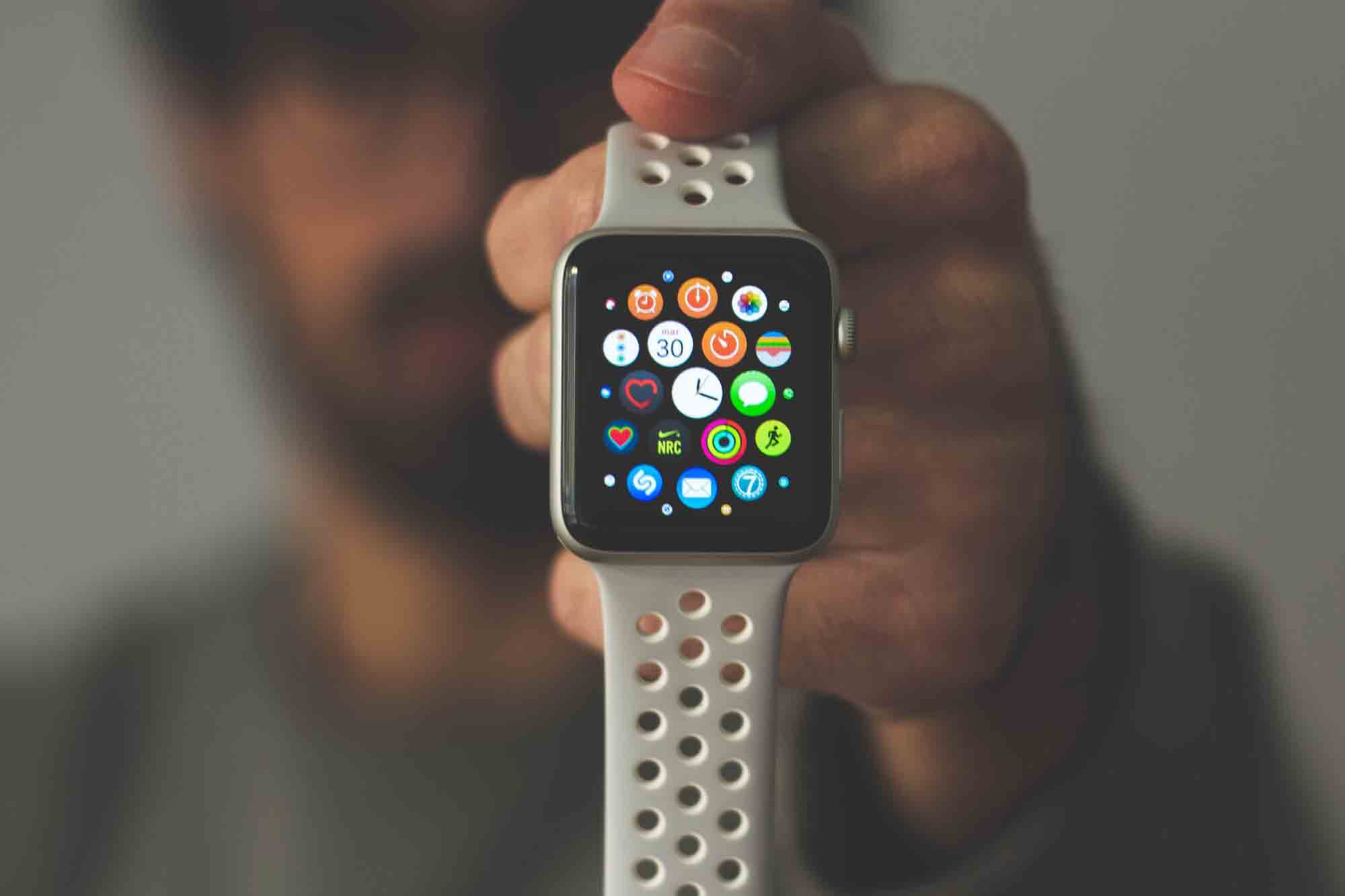 Male holding silver aluminum Apple Watch displaying apps with white Nike Sport Band, vertically in one hand