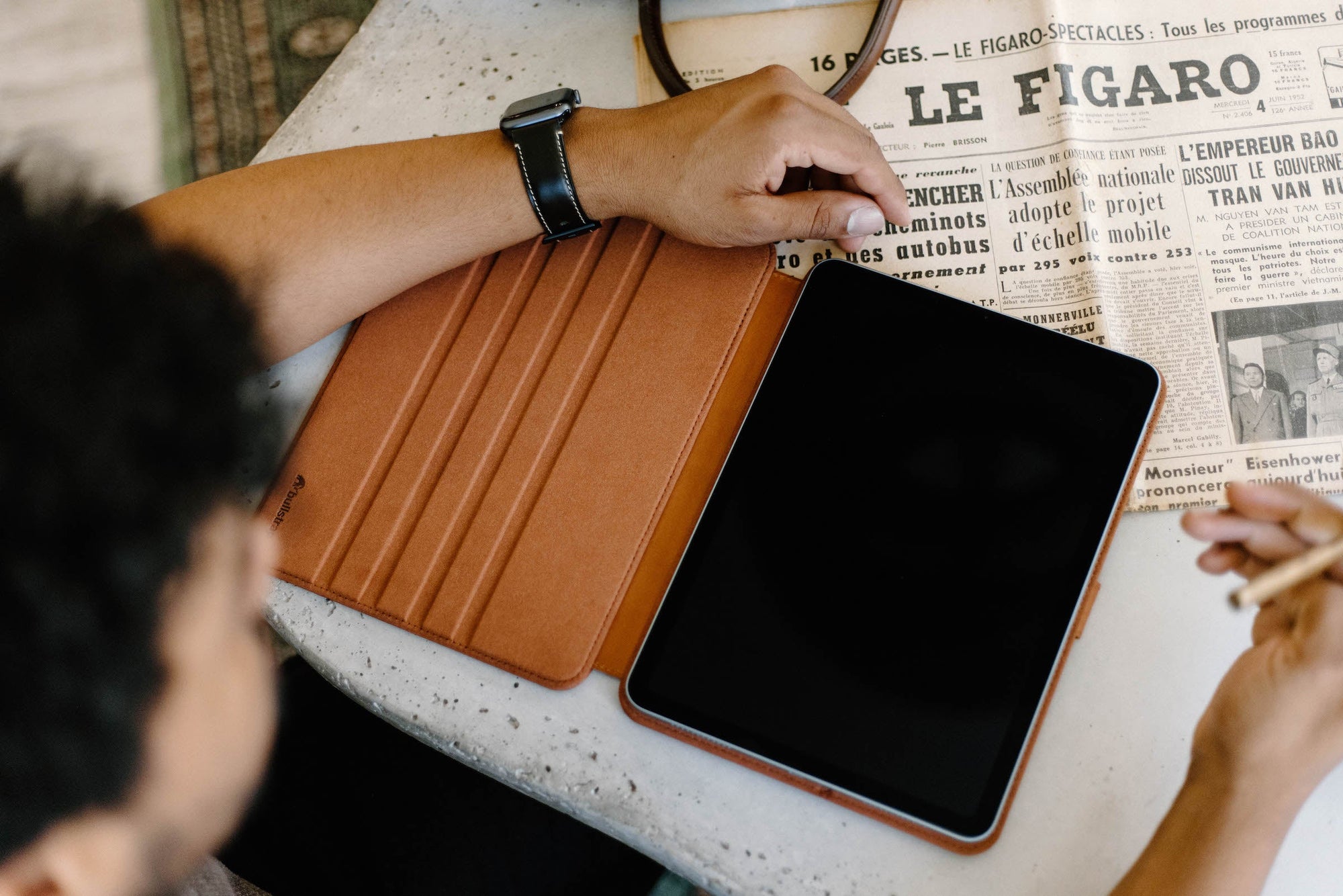 A person using a leather iPad case on a table with a newspaper.