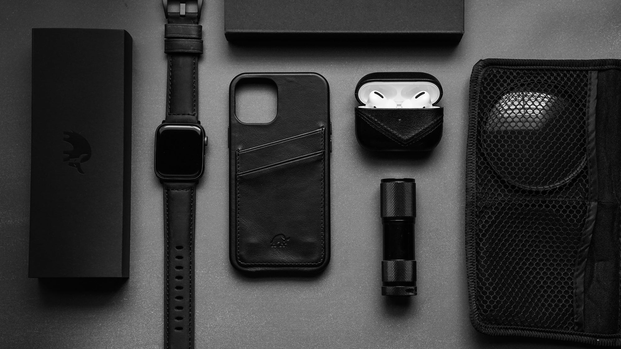 Black flat-lay everyday carry: leather phone card wallet, smartwatch, AirPods in leather case, compact flashlight.
