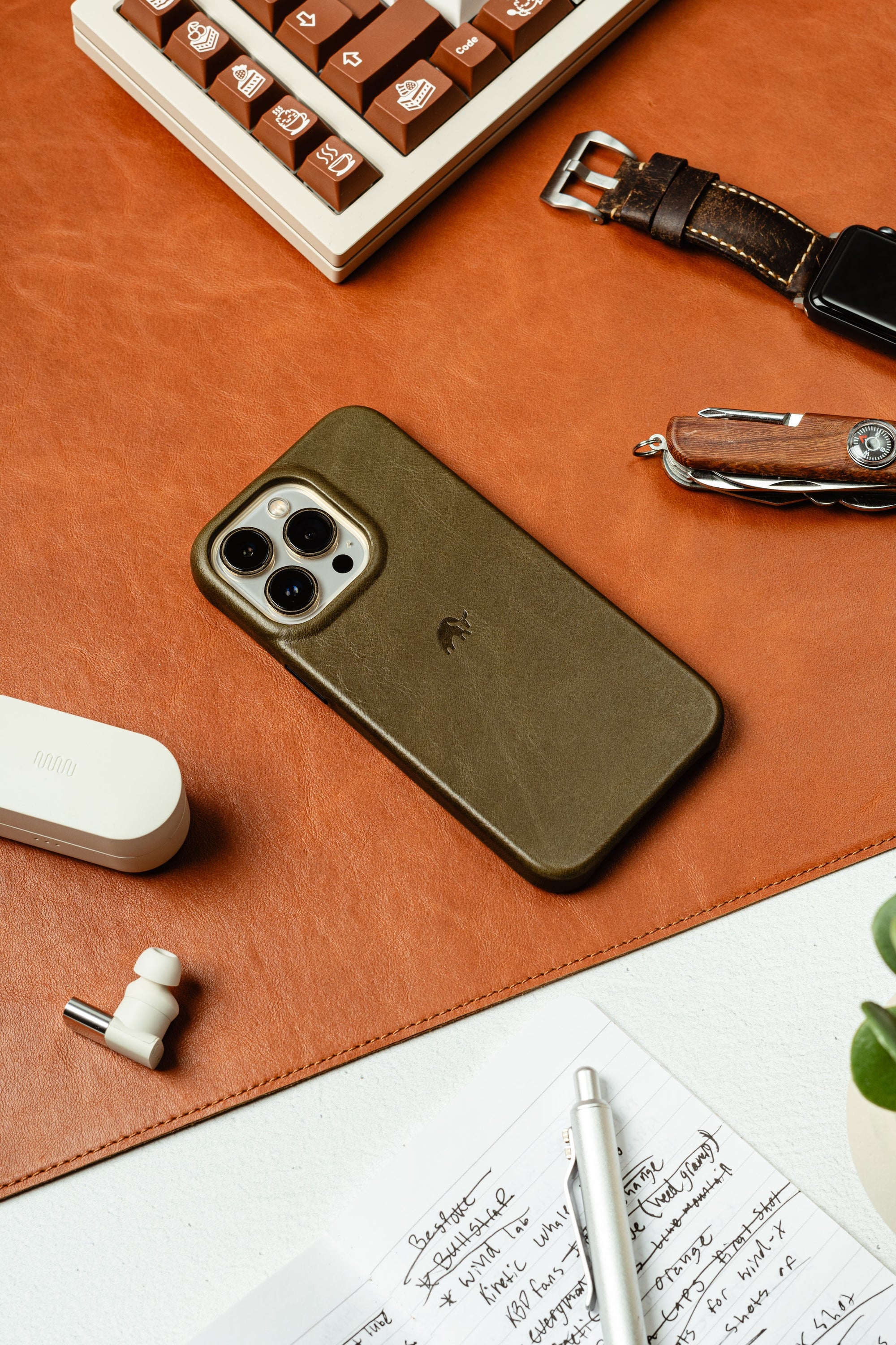 Olive green iPhone case on a desk with a keyboard and notebook