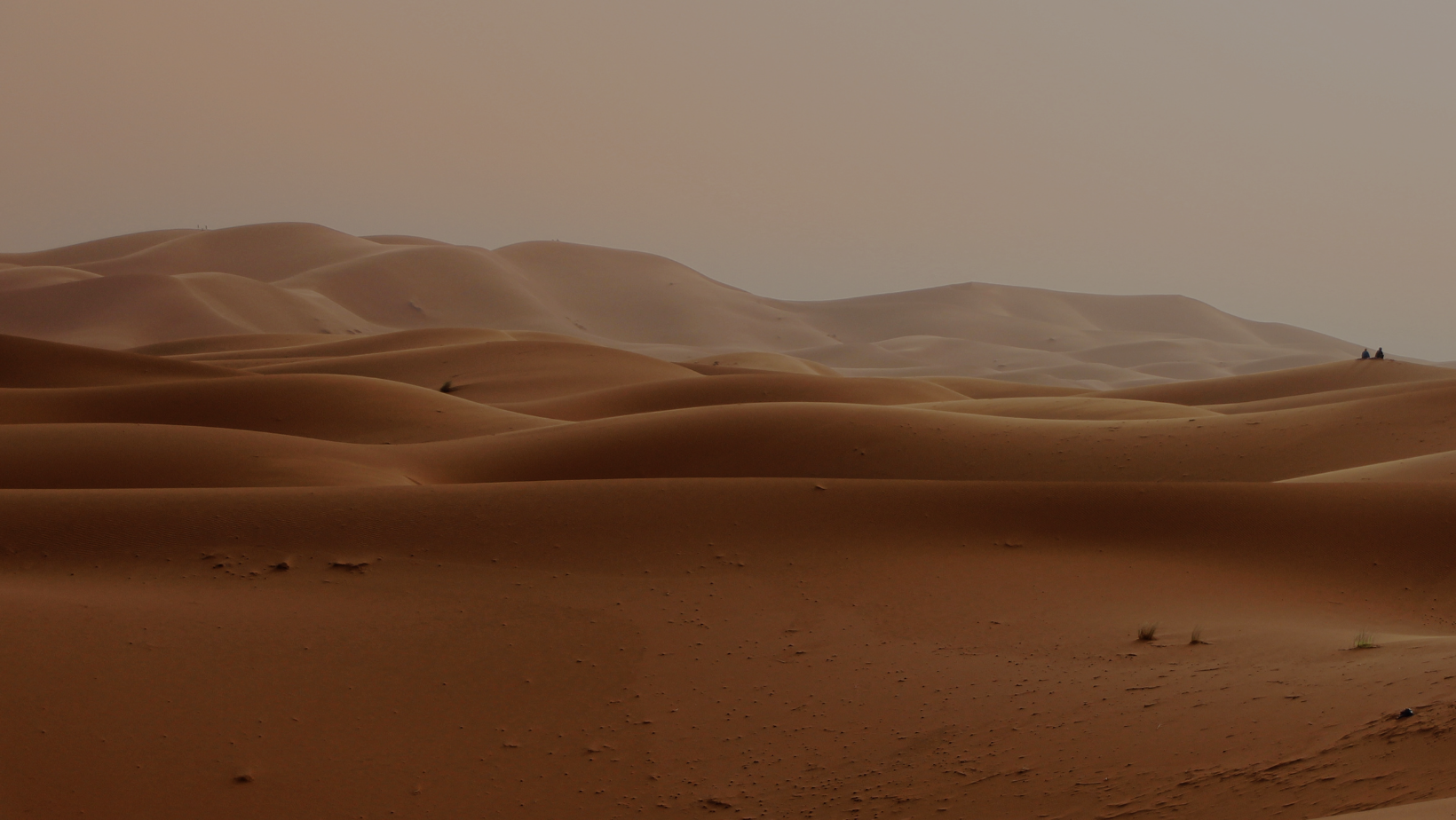 Desert dunes under a hazy sky with two people sitting on a distant dune