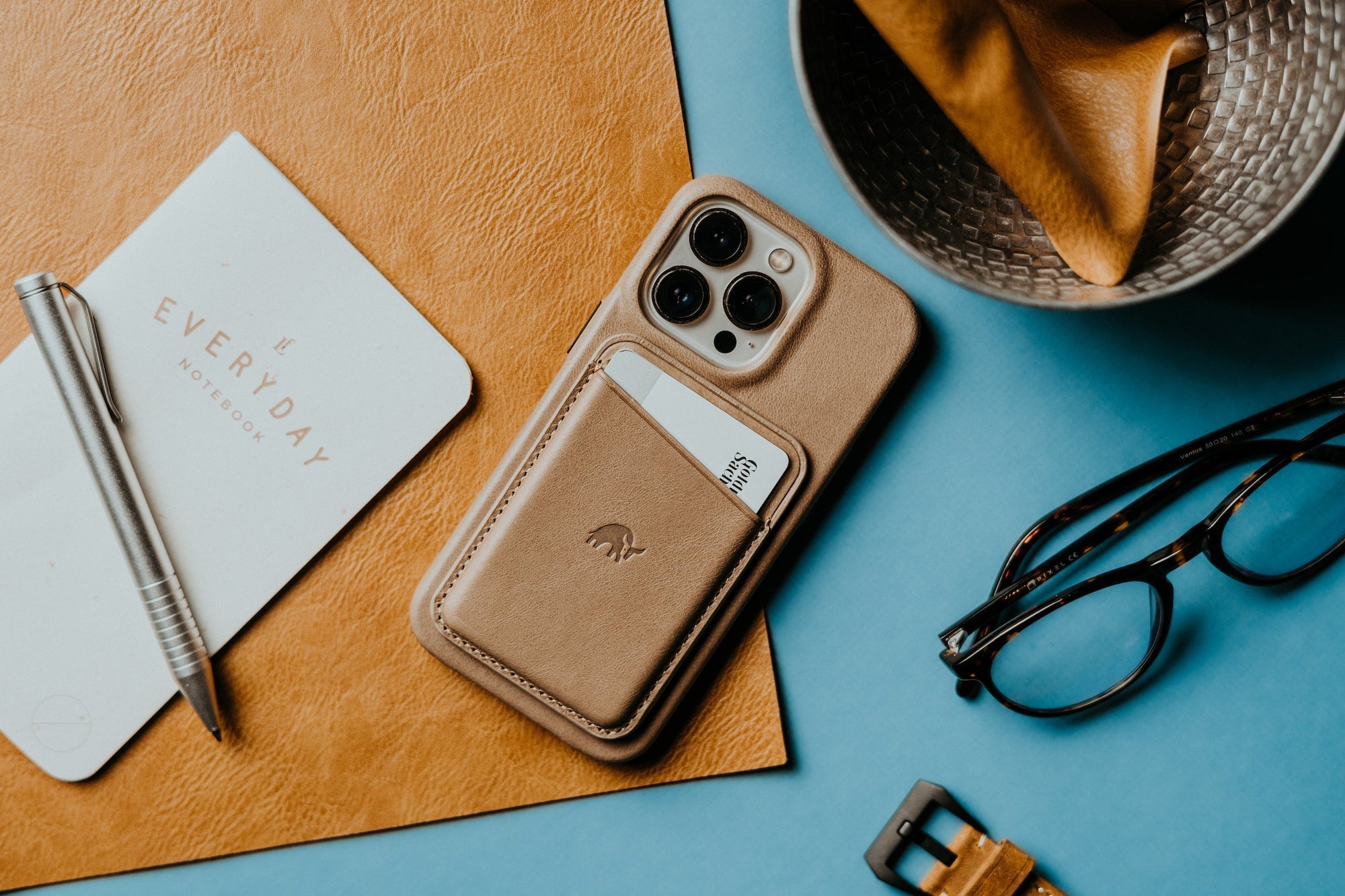 Smartphone with tan leather card wallet beside pen, EVERYDAY NOTEBOOK, glasses, and hat on blue and tan surface