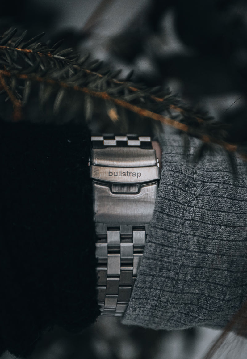 Stainless steel watch bracelet clasp engraved bullstrap, worn between dark jacket and gray sweater sleeves