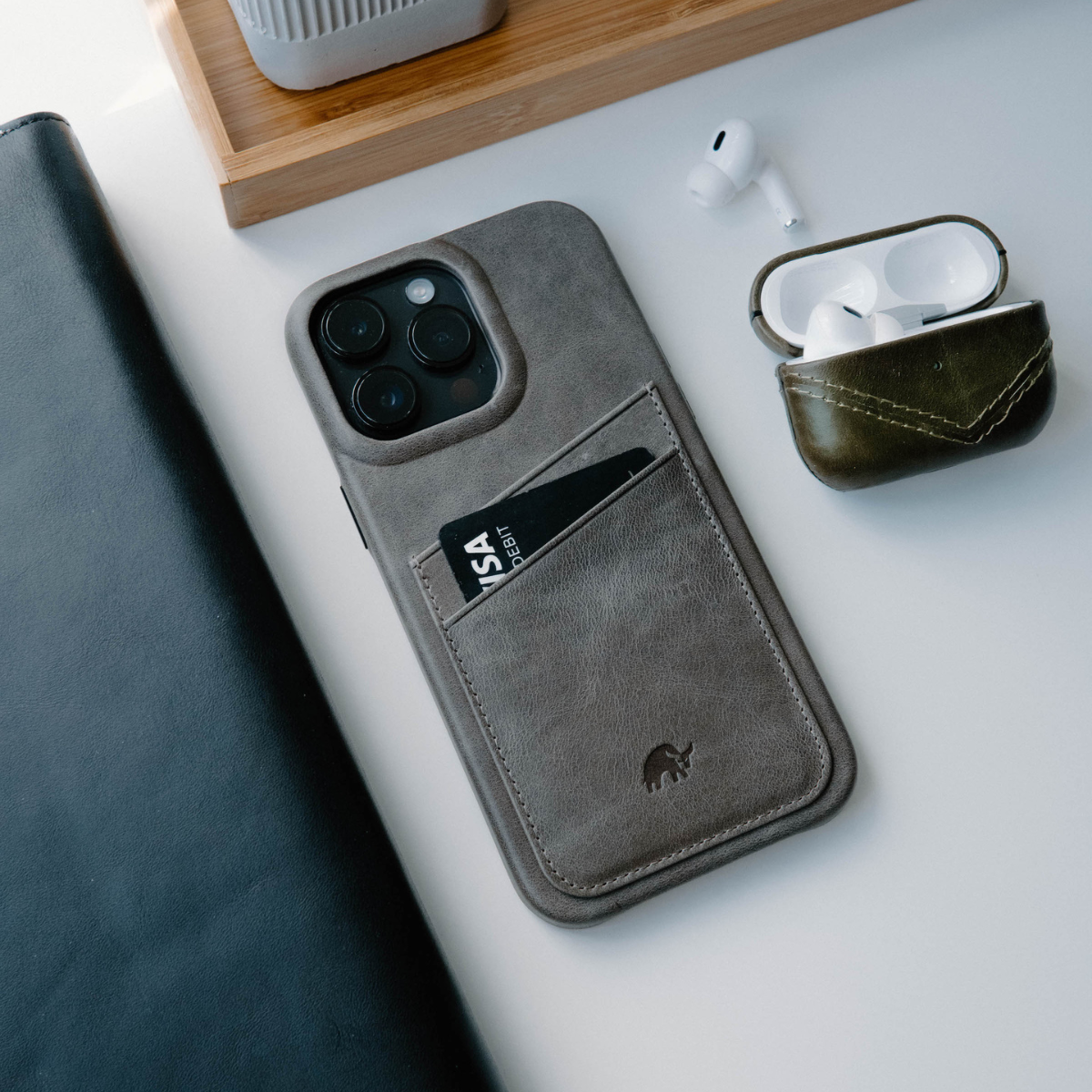 Slate minimalist phone wallet with a card labeled 'VISA DEBIT', beside open white AirPods and a black leather notebook.