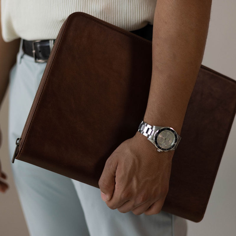 Close-up of brown leather MacBook sleeve held at hip showcasing smooth leather surface and zip closure against light clothing