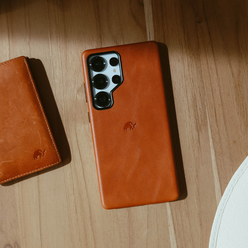 Brown leather phone case with multi-lens camera module on a wooden table, small matching leather wallet partly visible
