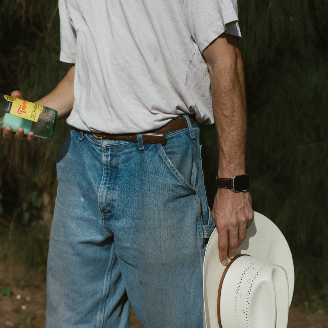 Man's torso in white T-shirt and jeans holding a green bottle labeled Topo in left hand and a white cowboy hat in right hand.