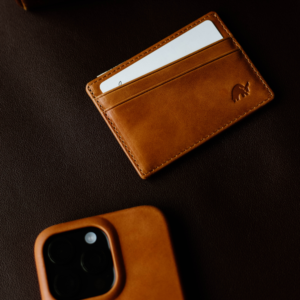 Brown leather card wallet with white card in pocket on dark leather surface; tan leather phone case visible lower-left.