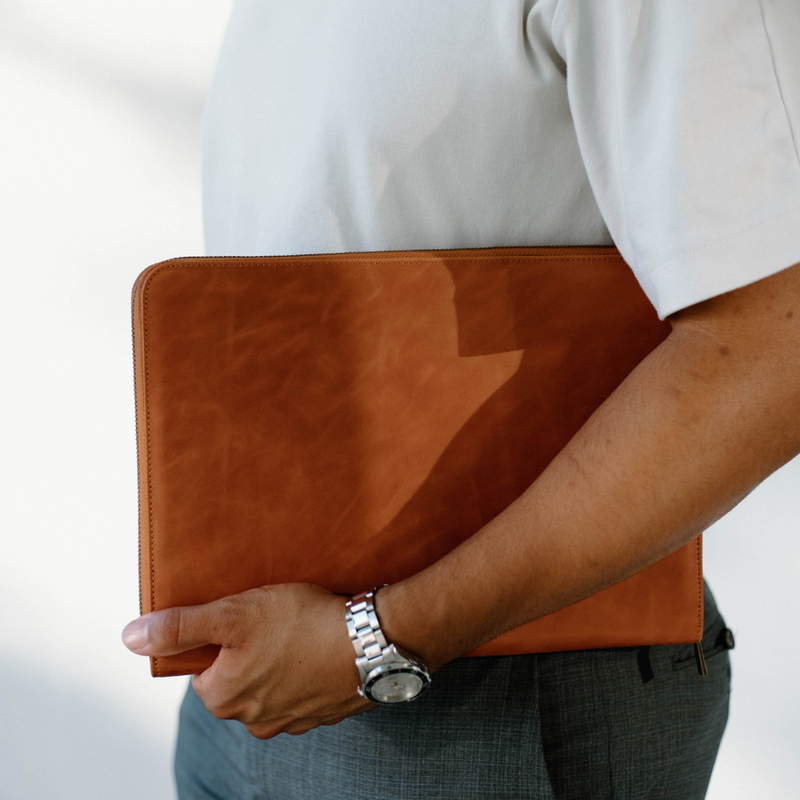 Person holding sienna leather laptop sleeve under arm against a neutral background, showing slim profile and zipper trim