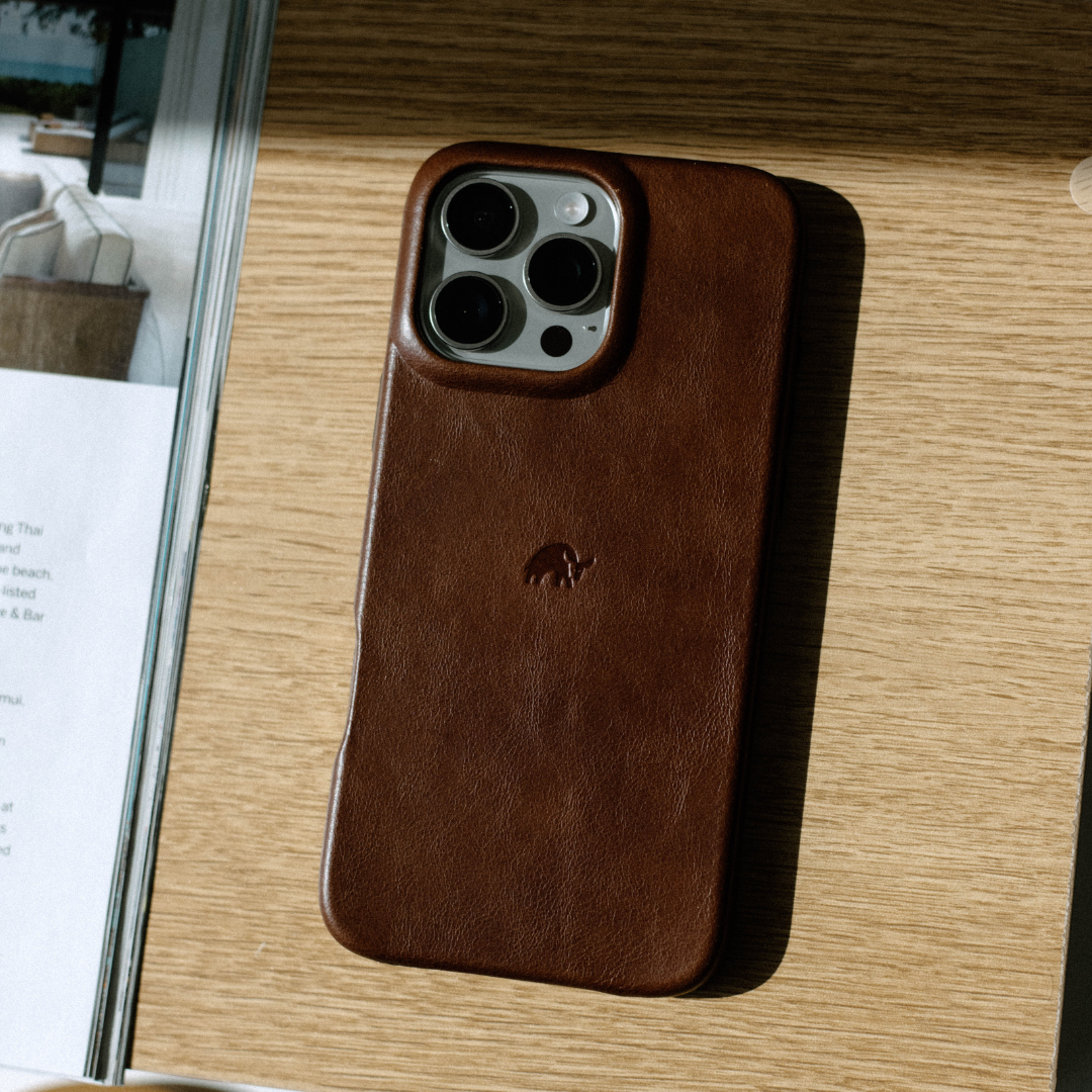 Brown leather phone case with raised camera frame and embossed animal logo on a light wood table beside a magazine.
