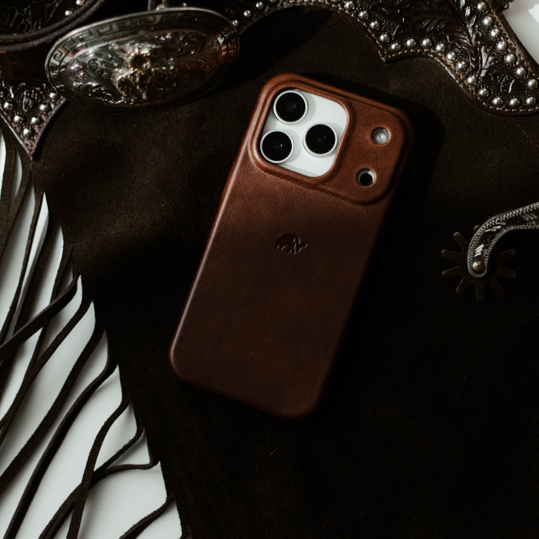 Brown leather phone case (TERRA) on dark fringed fabric, showing triple-lens camera cutout