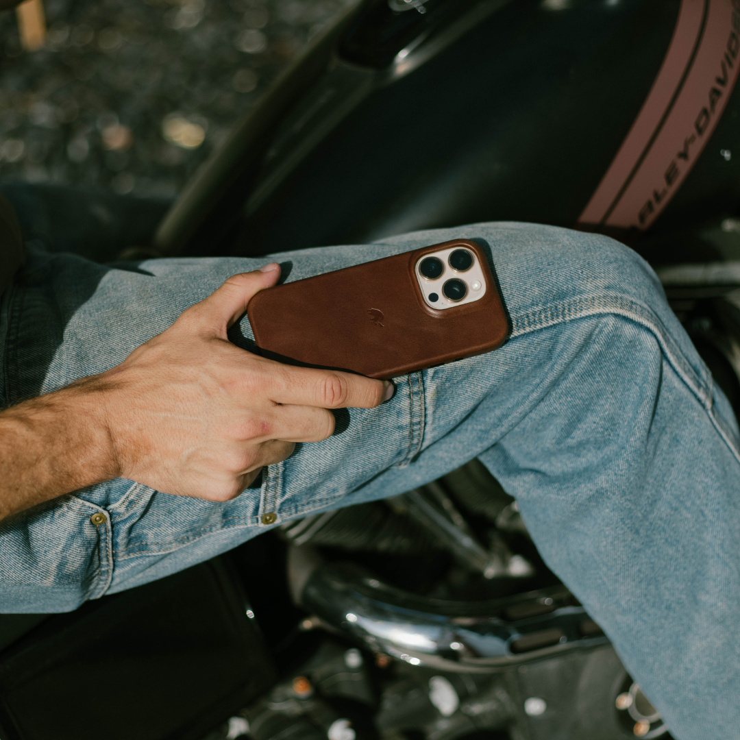 Brown leather iPhone case held on a person's denim-clad lap, phone resting on a motorcycle seat
