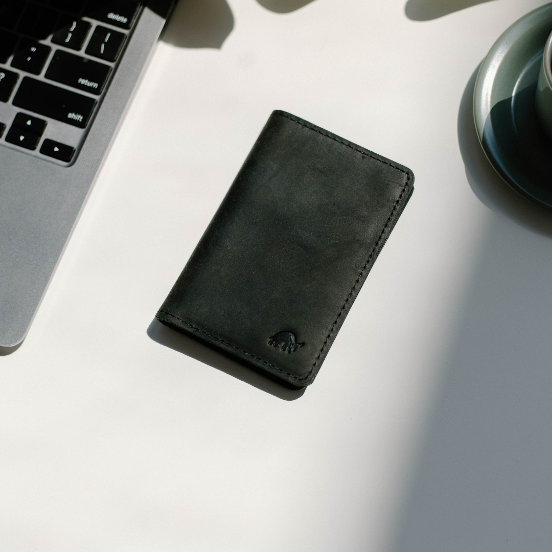 Black leather vertical wallet on white desk beside laptop keys labeled return, shift, delete, and a cup on a saucer.