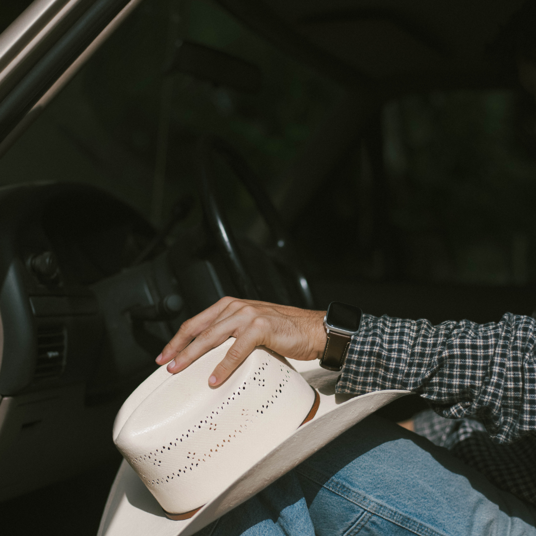 Hand with smartwatch resting on a white perforated cowboy hat on a lap inside a car.