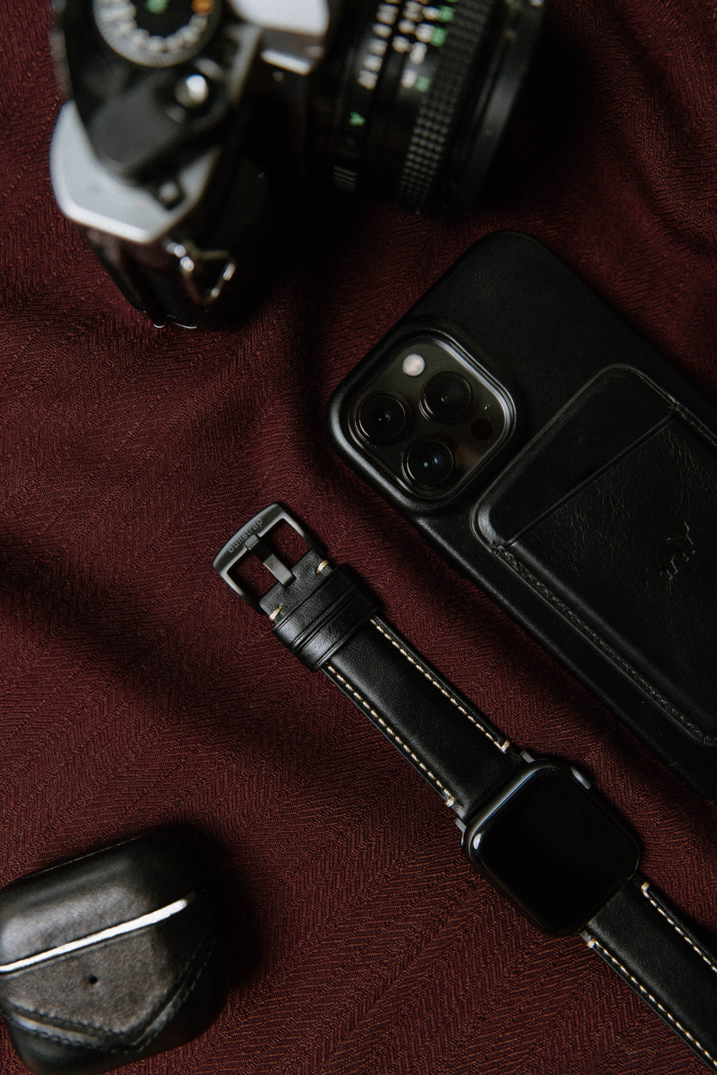 A leather watch strap next to a camera, phone, and AirPods on a maroon fabric background.