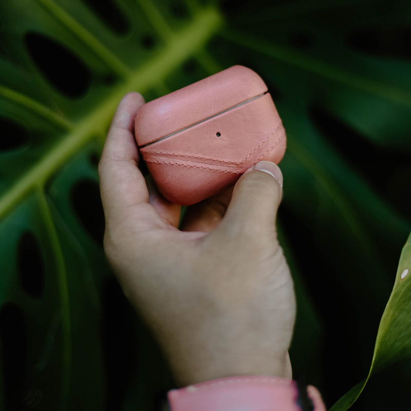 Hand holding coral leather AirPods case with stitched chevron detail against large green leaf background