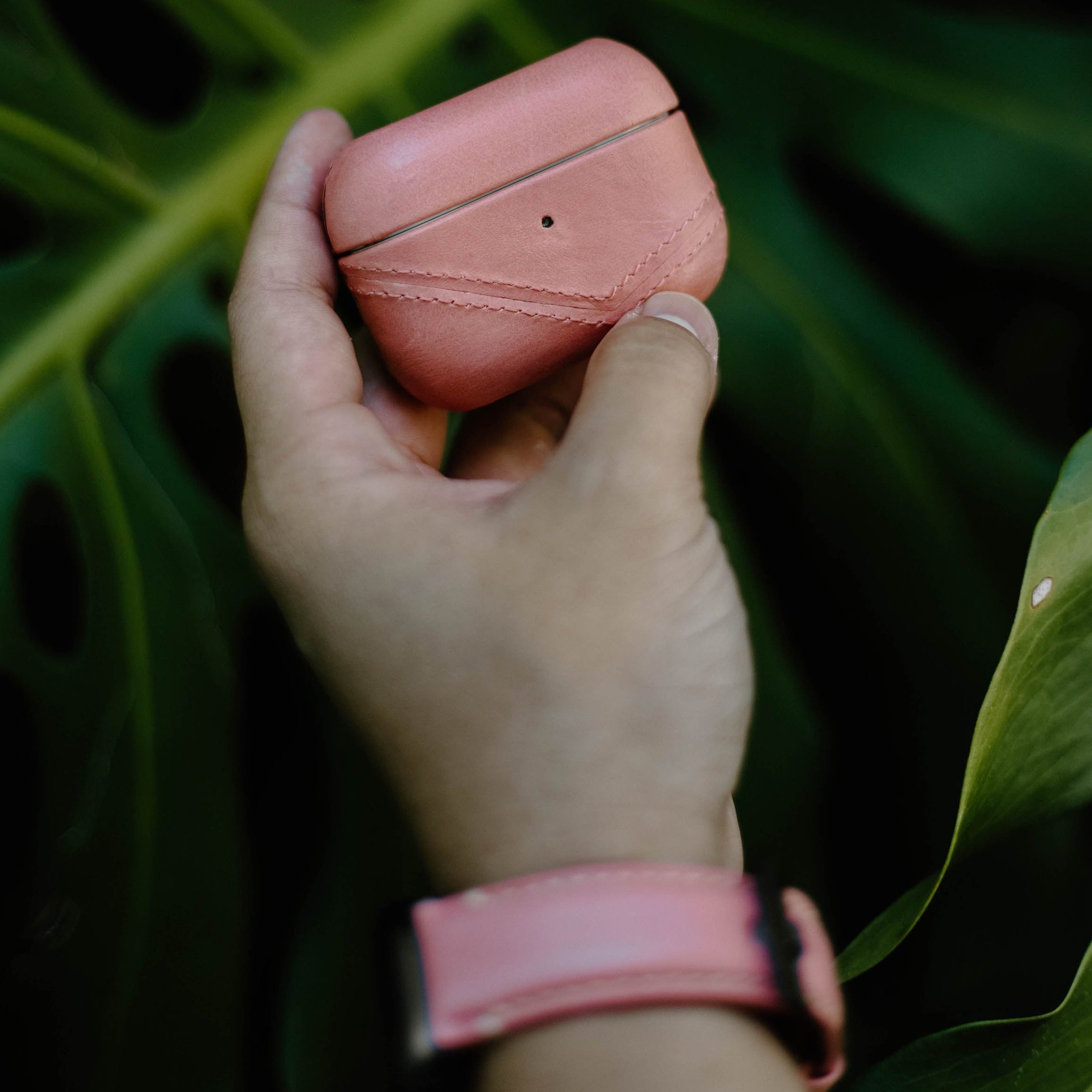 Hand holding coral leather earbud case with matching coral leather Apple Watch strap on wrist against tropical leaf background