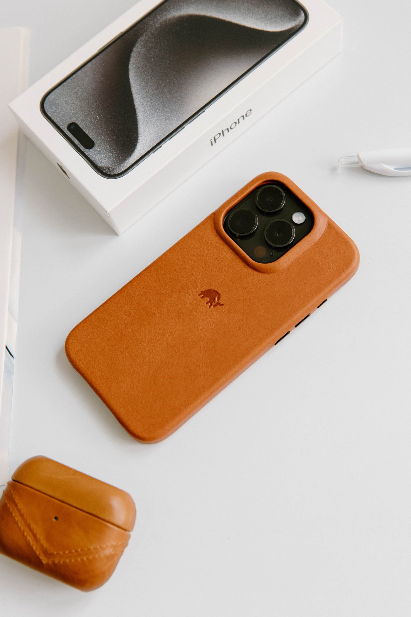 A leather iPhone case in brown color with an iPhone and AirPods on a white surface.