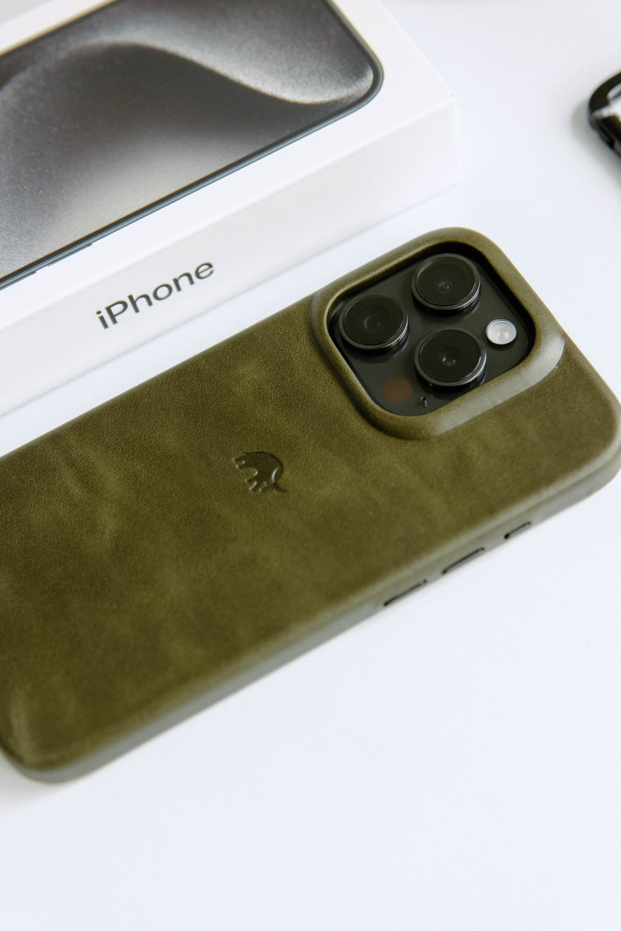 An olive green leather iPhone case with camera cutouts next to an iPhone box.