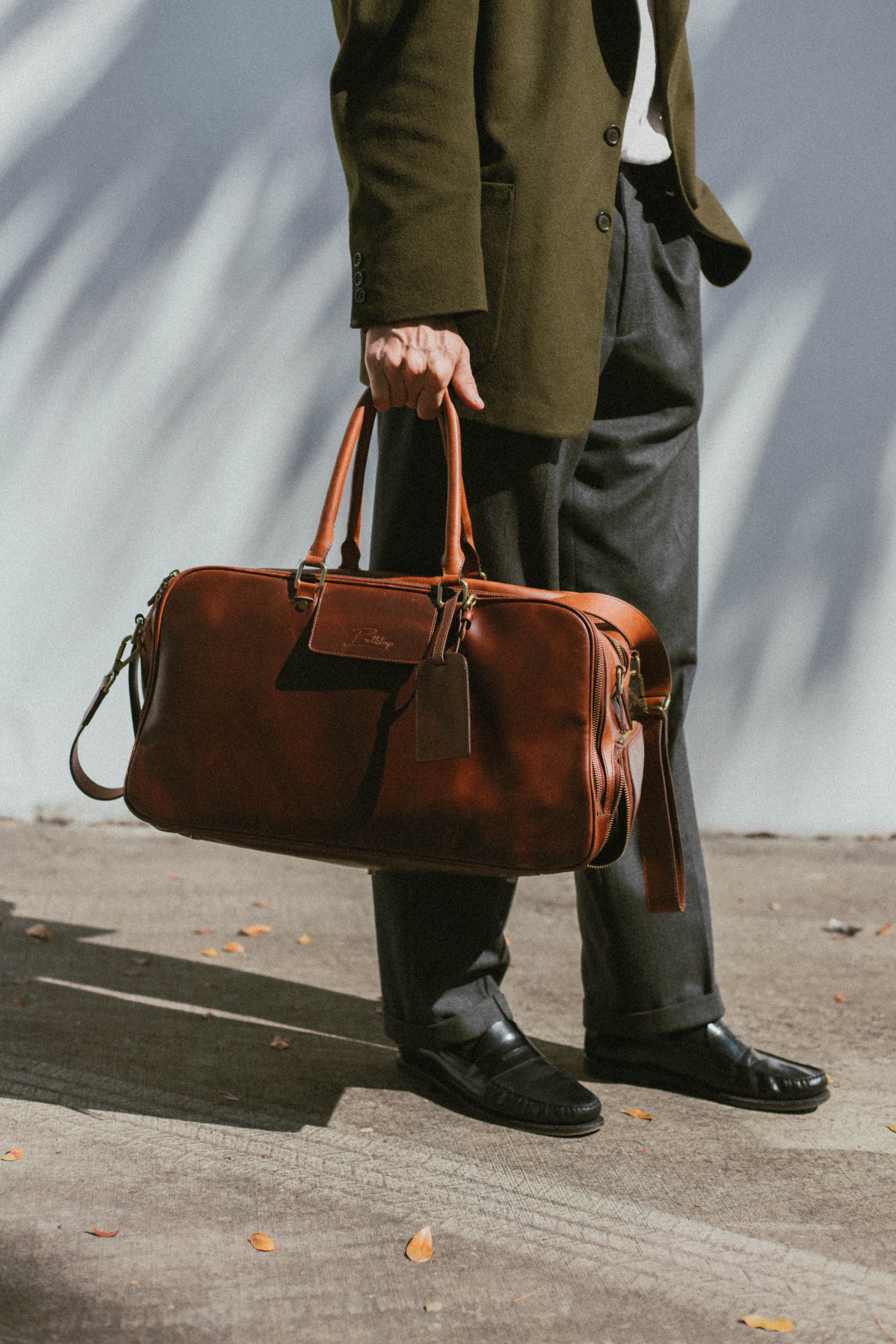 Person holding brown leather weekender bag by the handles, wearing olive coat, gray trousers and black loafers on sunlit pavement.