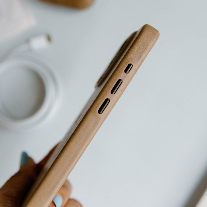 Side view of a tan leather phone case showing three cutouts for buttons, with white charging cable blurred in background.