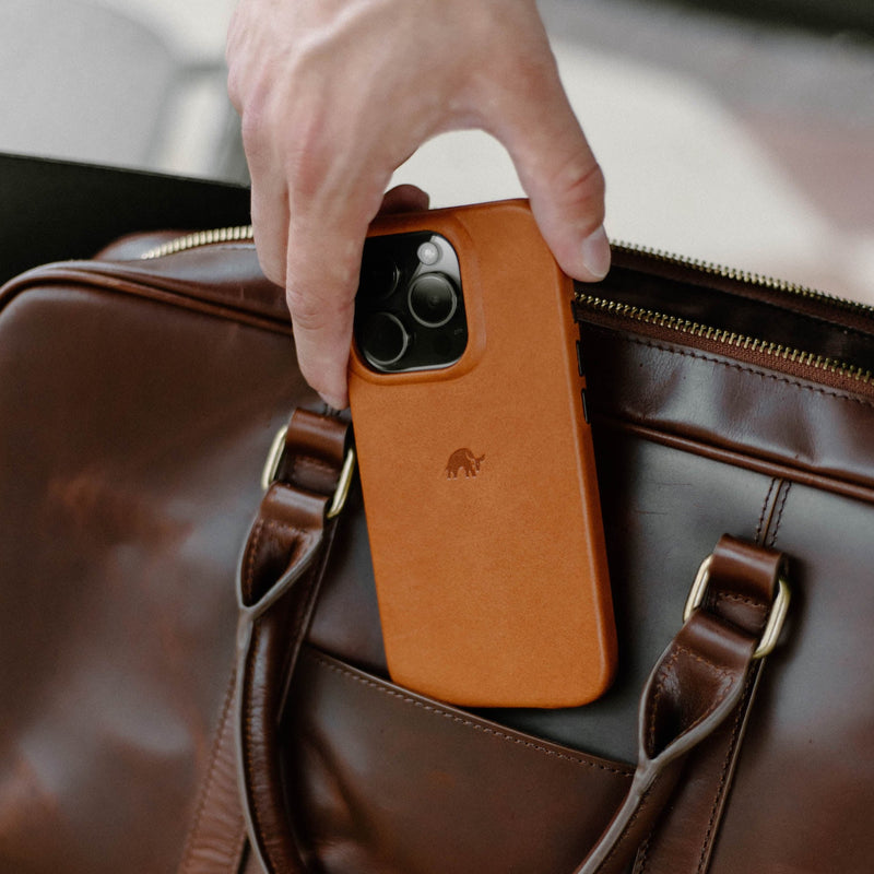 A hand holding a leather phone case above a brown leather bag.