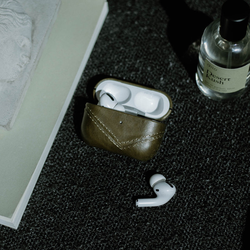 A leather AirPods case in brown color with AirPods inside, placed on a textured surface next to a book and a bottle.