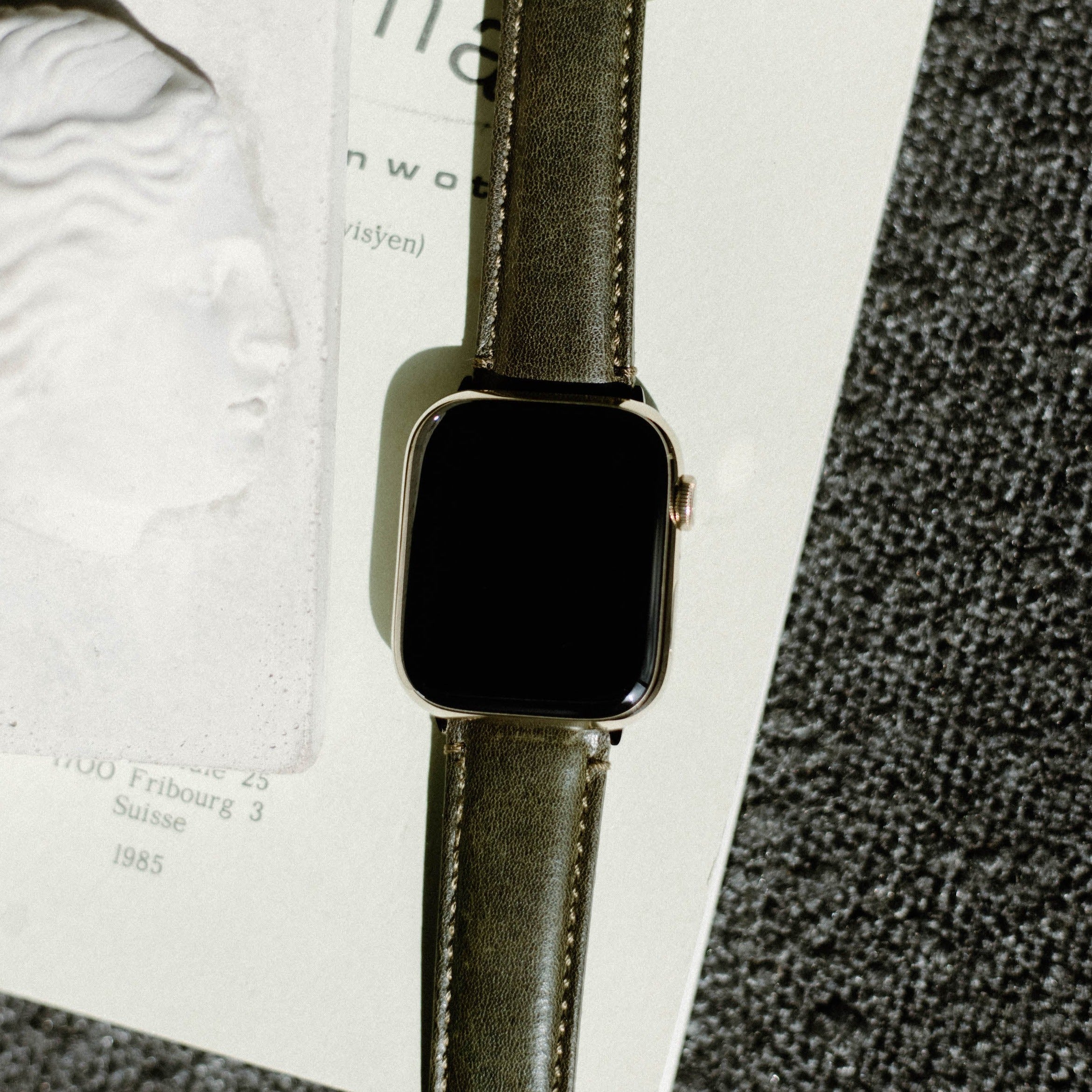 Olive-green leather Apple Watch strap (Maverick) with blank black face on page showing 'Fribourg 3 Suisse 1985'