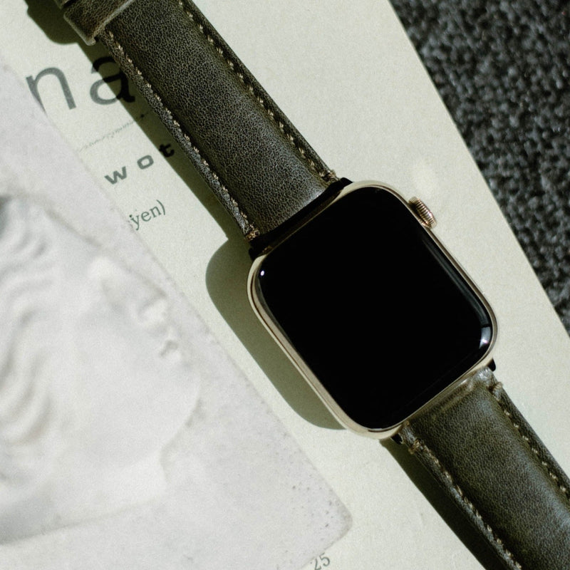 Angled top-down view of Apple Watch with Maverick olive leather band showcasing pebble-grain texture and contrast stitching on a light paper background
