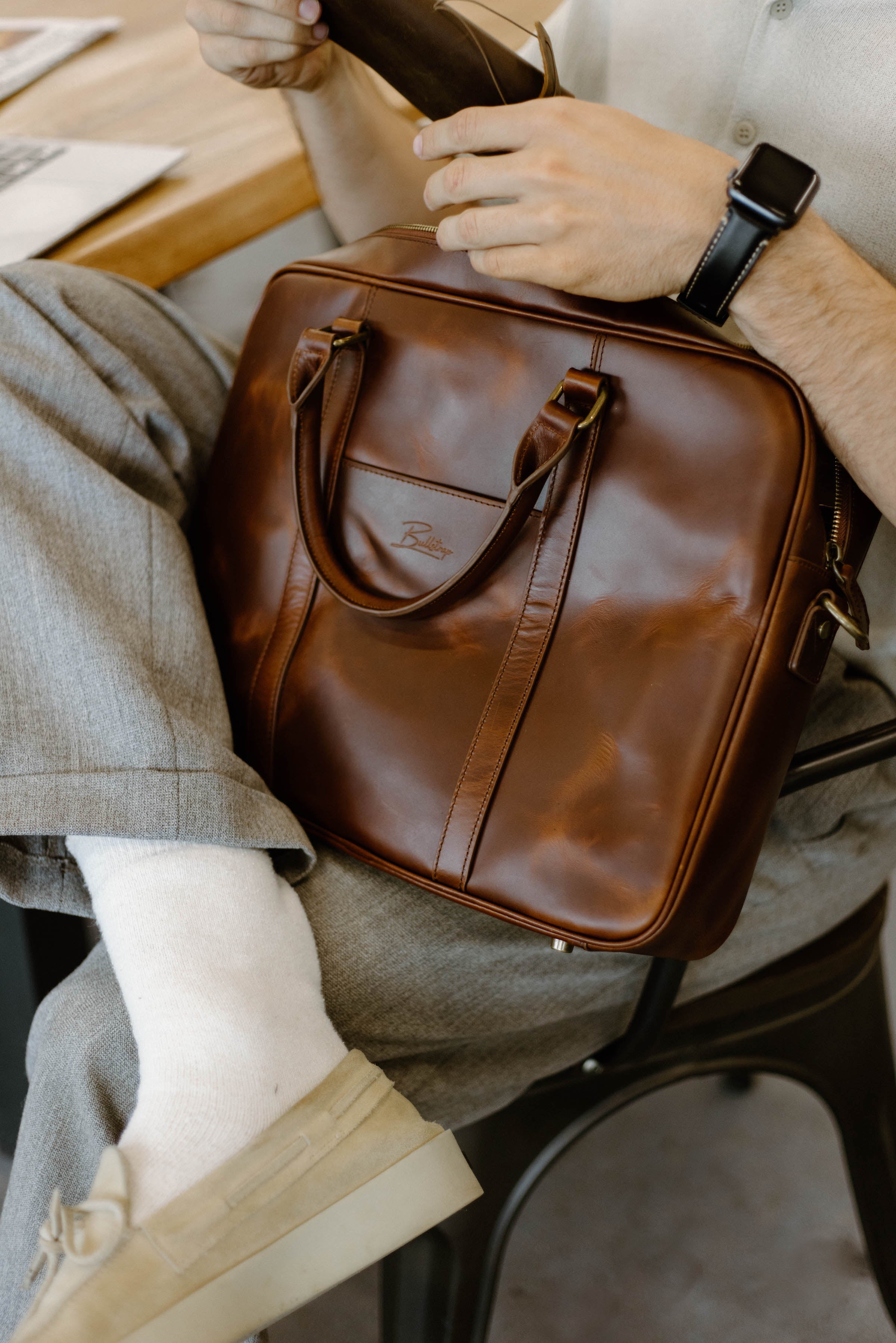 Brown leather briefcase with embossed Bullstrap logo; close-up showing handle and a hand on the zipper.