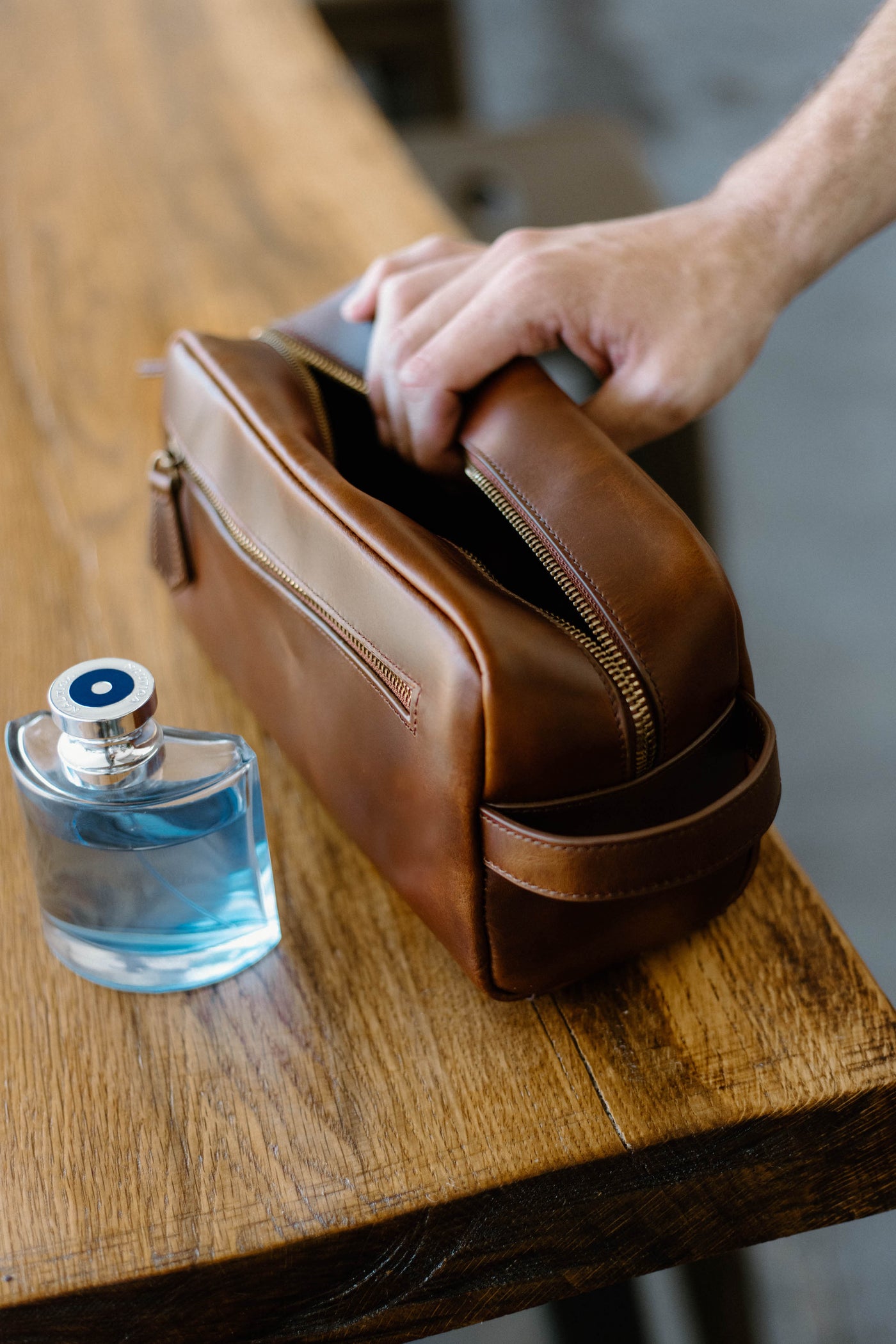 A leather toiletry bag next to a bottle of cologne on a wooden table.