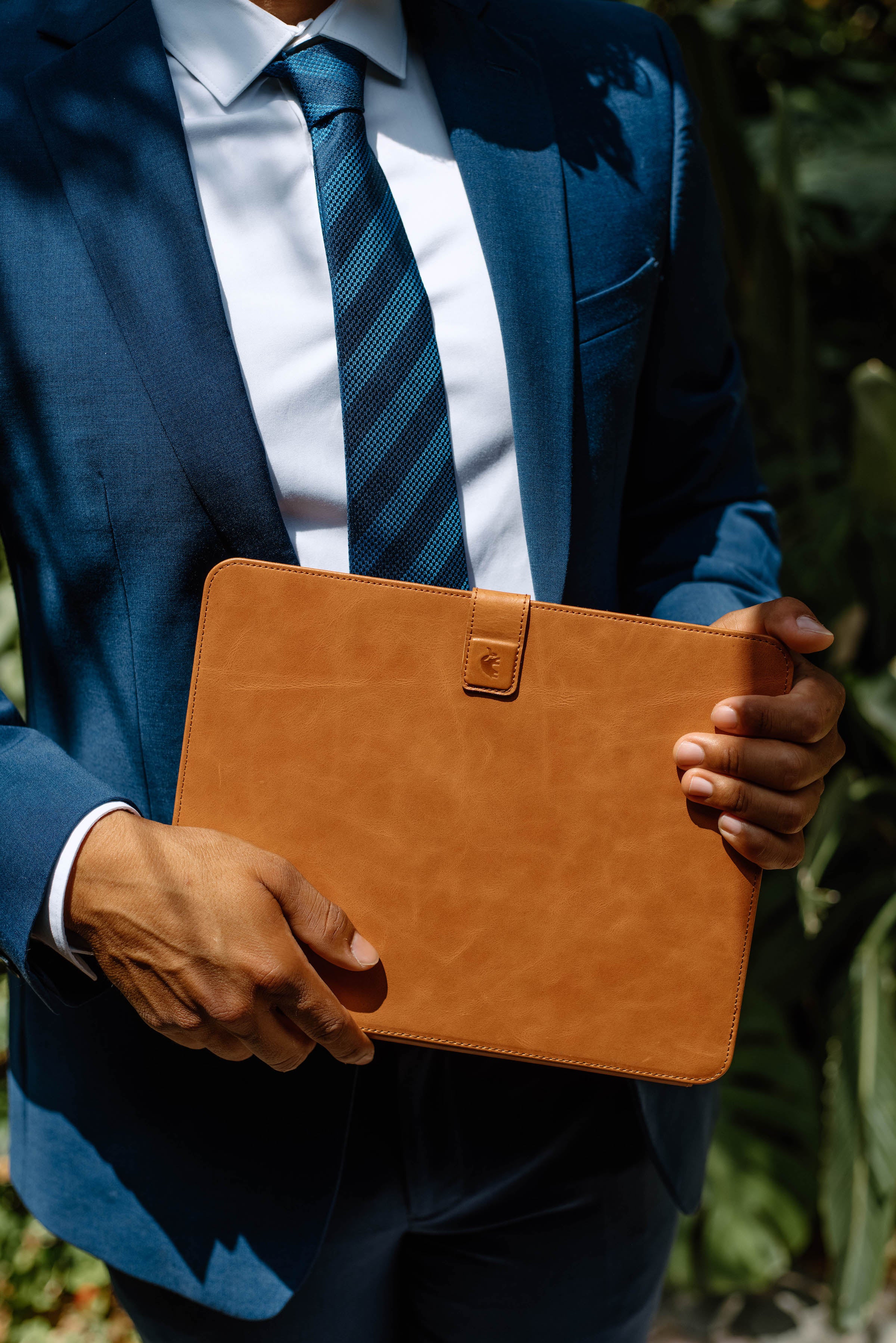 Person in blue suit with striped tie holding a tan leather tablet folio against their torso