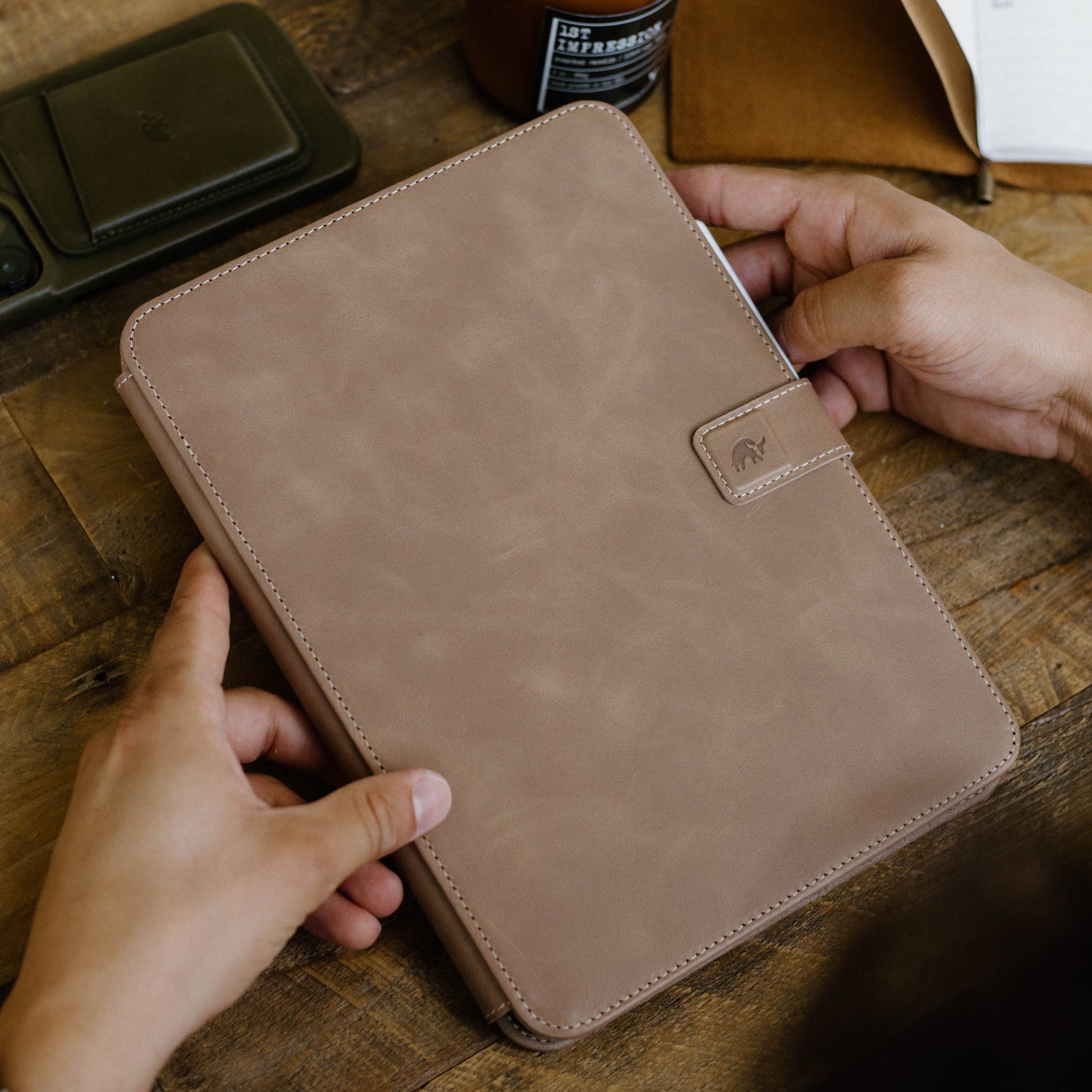 Tan DUNE leather iPad folio with elephant-embossed strap on a wooden table; bottle label reads 1ST IMPRESSION.