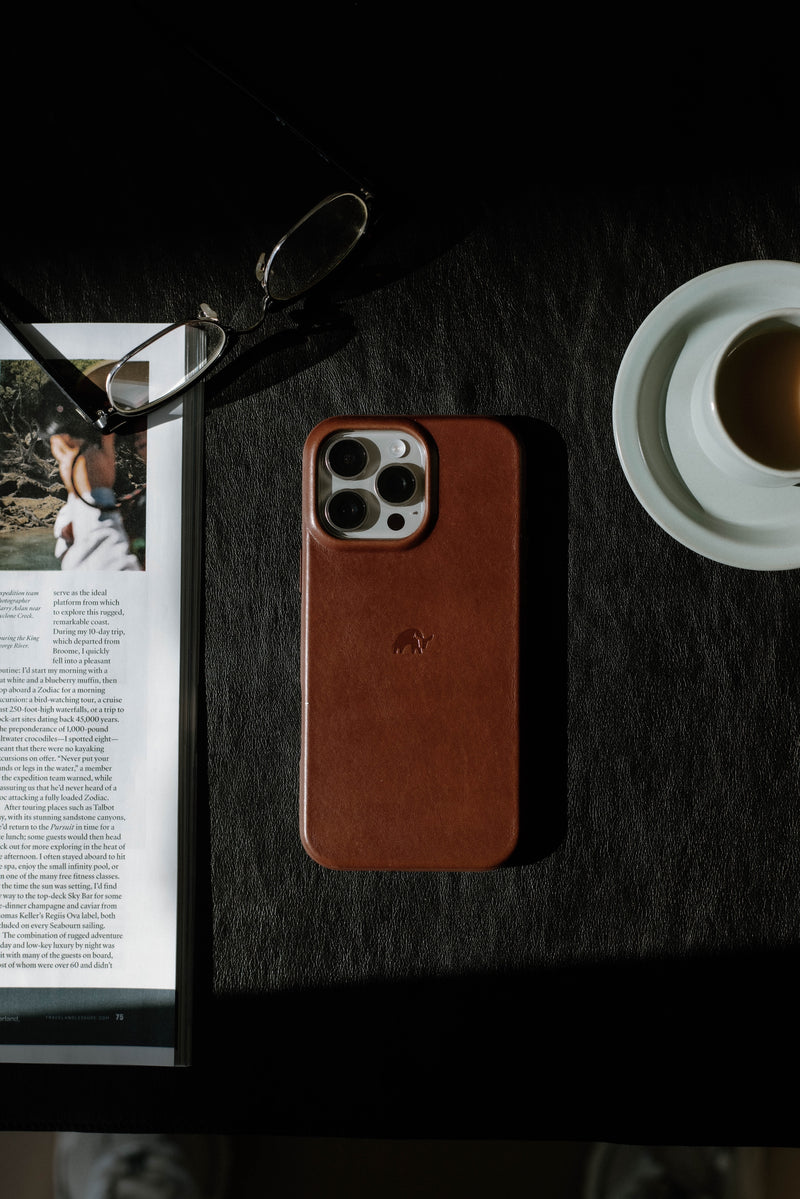 A brown leather phone case on a table next to a magazine and a cup of coffee.