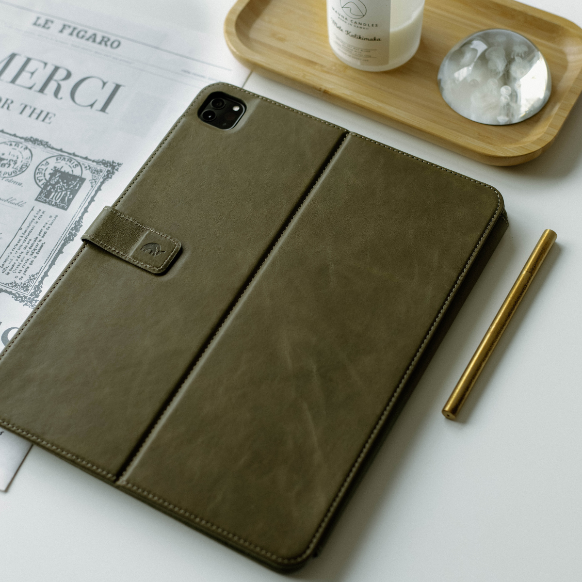 Olive green leather iPad case with a flap and embossed logo on a white background with a pen and decorative items.