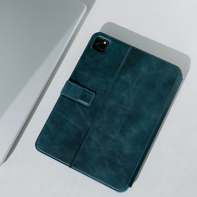 Teal leather folio tablet case with camera cutout and magnetic strap, photographed beside a silver laptop.