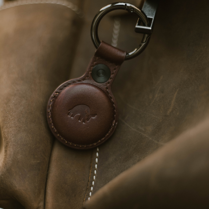 Brown leather key fob with embossed elephant logo attached to a metal key ring, resting on a stitched leather bag.