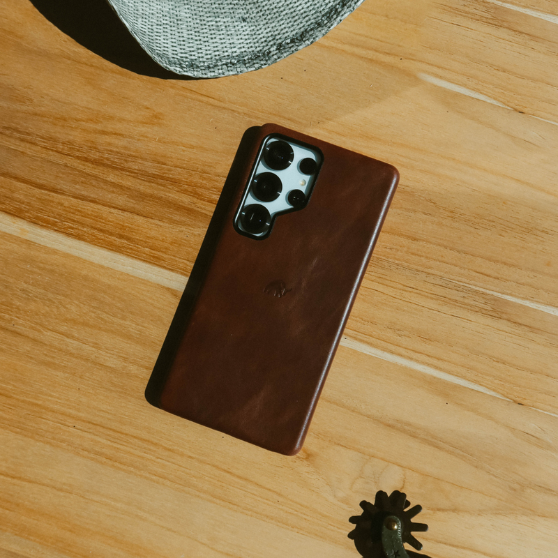 Brown leather phone case with visible camera lenses on wooden tabletop; straw hat edge top left and small spur bottom right.