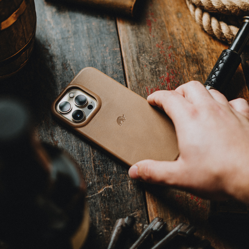 Hand reaching for iPhone with brown leather case on a worn wooden table, with coiled rope and tools nearby.