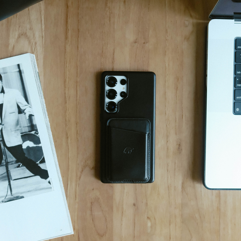 Black smartphone with leather card wallet on table between open magazine (page 19) and laptop keys esc tab cap shift