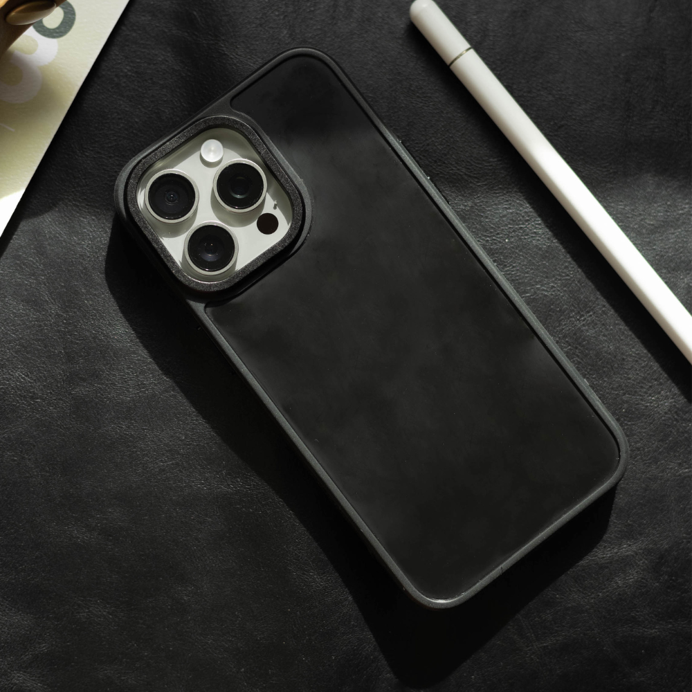 Black smartphone in a black case showing triple-lens camera, next to a white stylus on a dark leather surface.