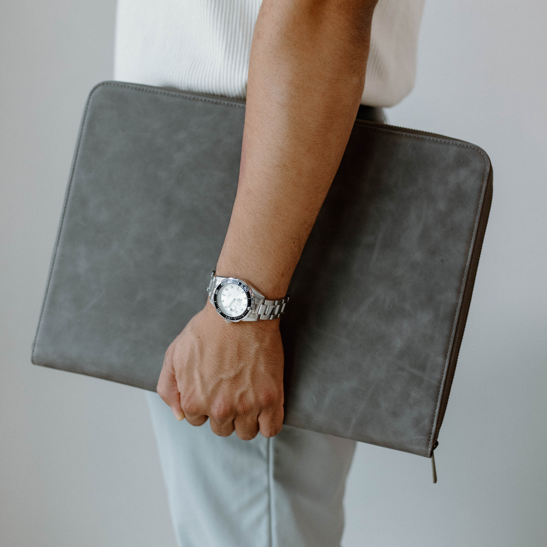 A person holding a slate-colored leather MacBook sleeve with a watch on their wrist.