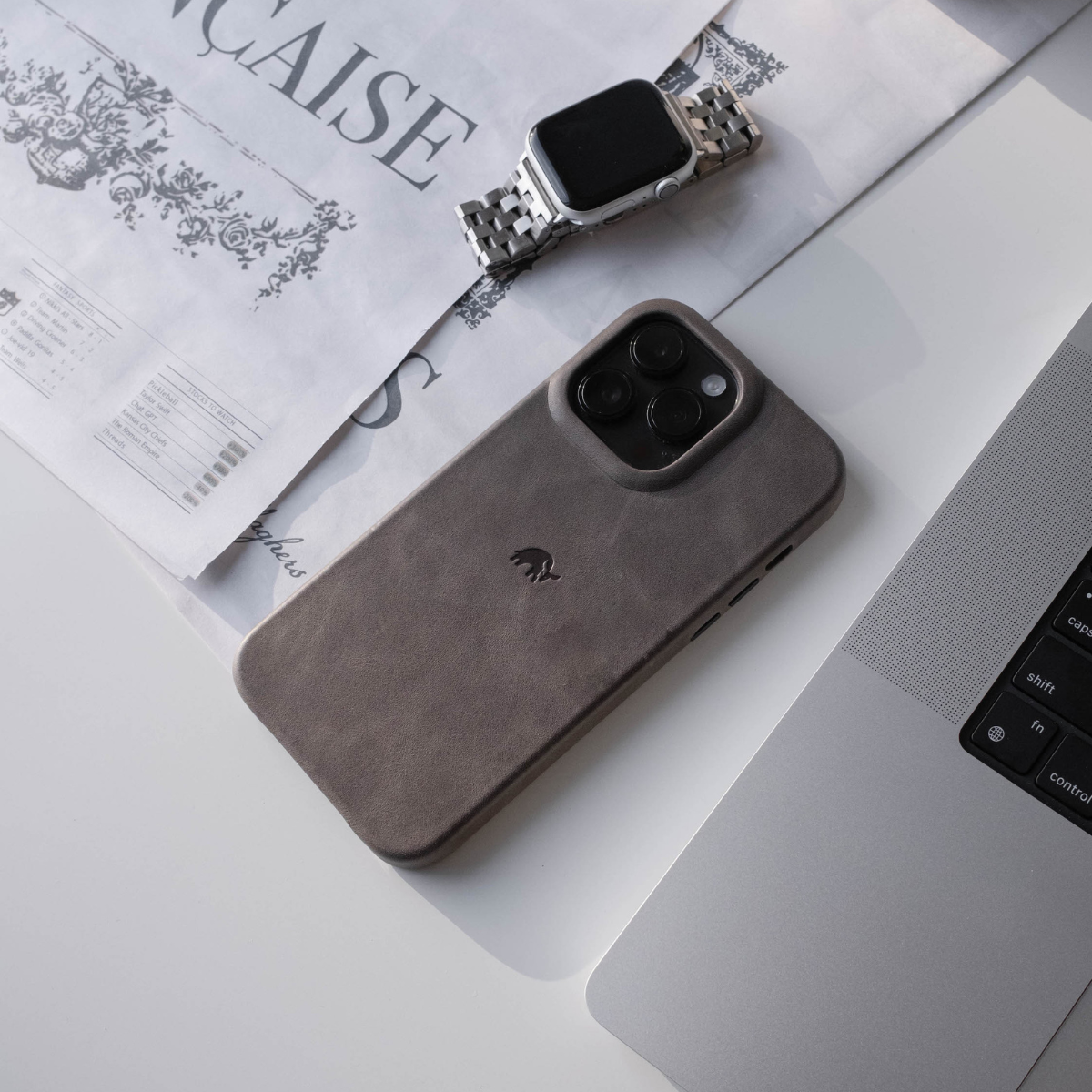 Phone with grey leather case and triple camera beside smartwatch and laptop keys fn control shift caps; words CAISE speakers