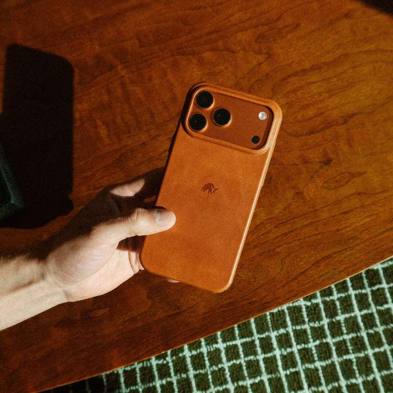 Hand holding Contemporary Plateau Case in Sienna showing full-grain leather and MagSafe camera cutout on wood table