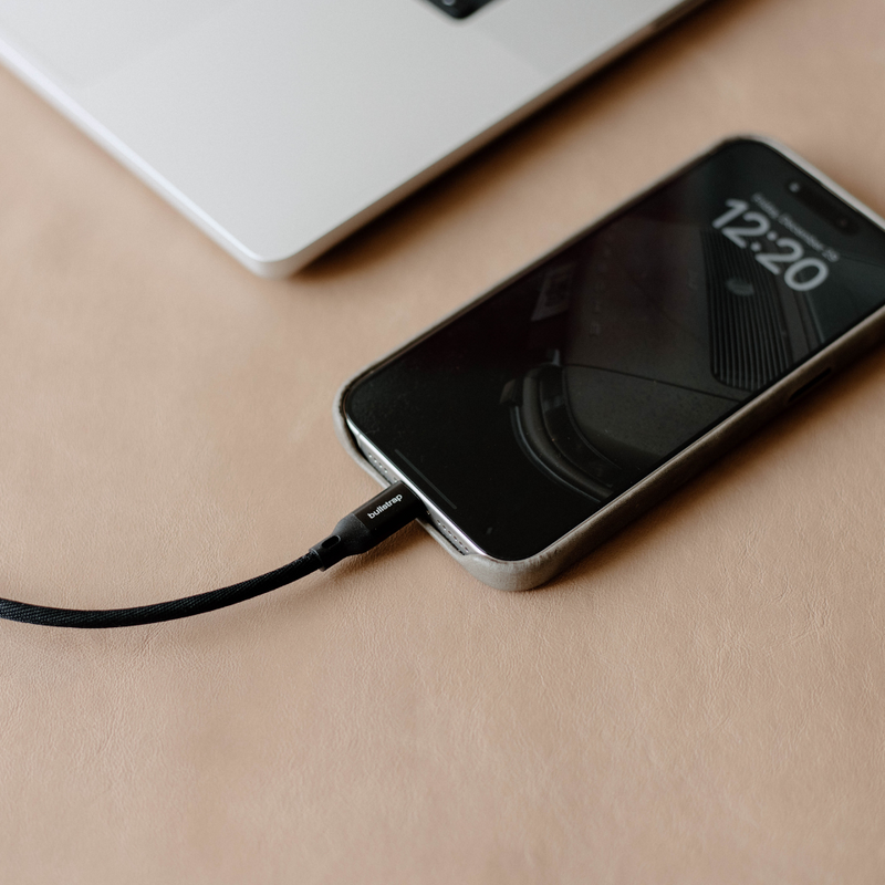 Close-up of Lightning charging cable plugged into an iPhone showing braided black cable and bullstrap-branded connector on tan leather surface