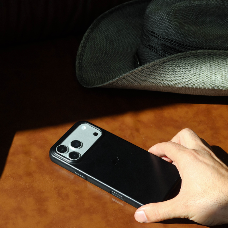 Three quarter view of Contemporary Plateau black leather MagSafe phone case showing raised camera lip and edge-to-edge leather, hand holding with hat in background