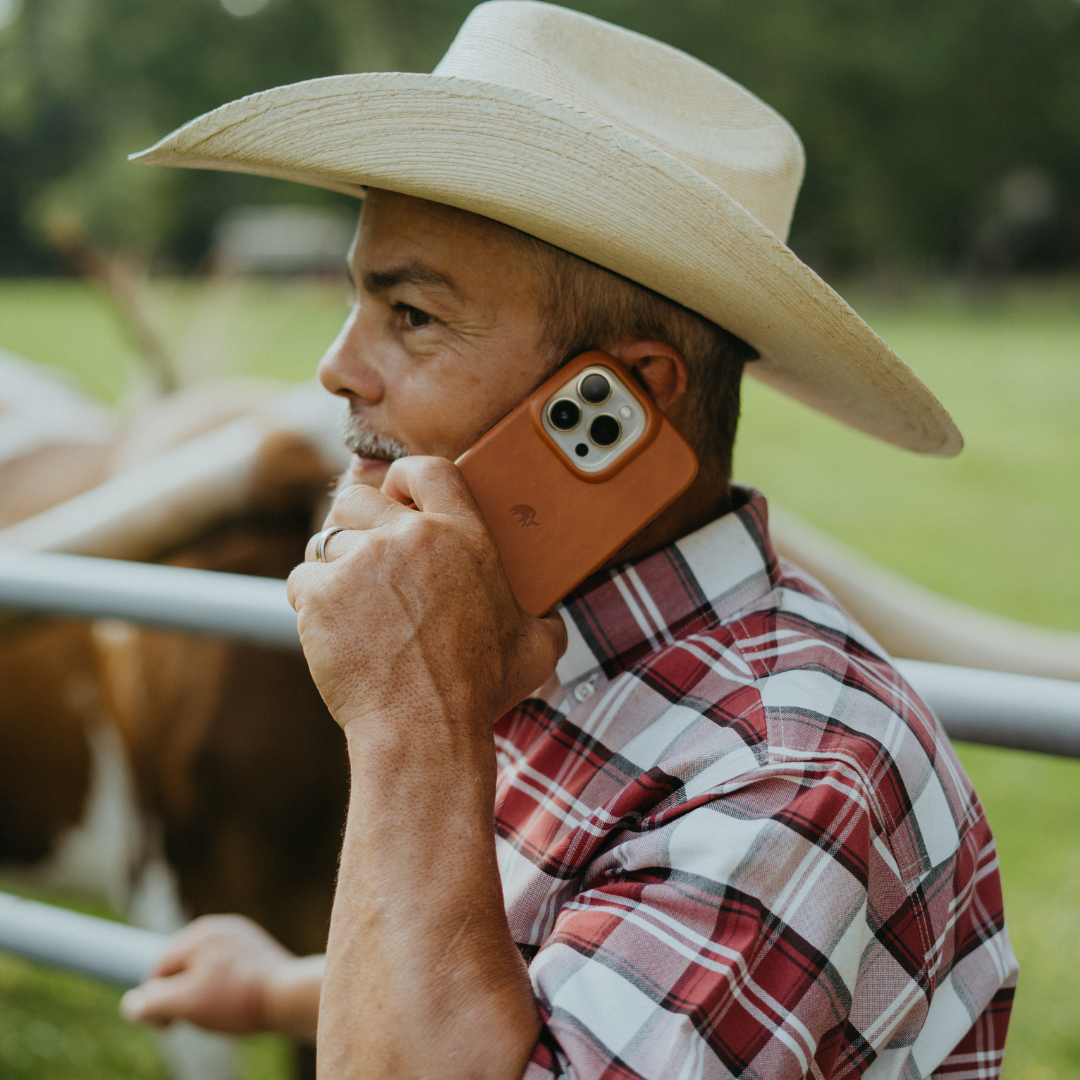 Cowboy holding iPhone in sienna leather case against a pasture, close-up profile view.