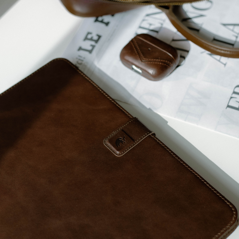 Angled top-down view of Terra brown leather iPad case showing stitched magnetic clasp and embossed logo on a white tabletop beside a matching leather AirPods case