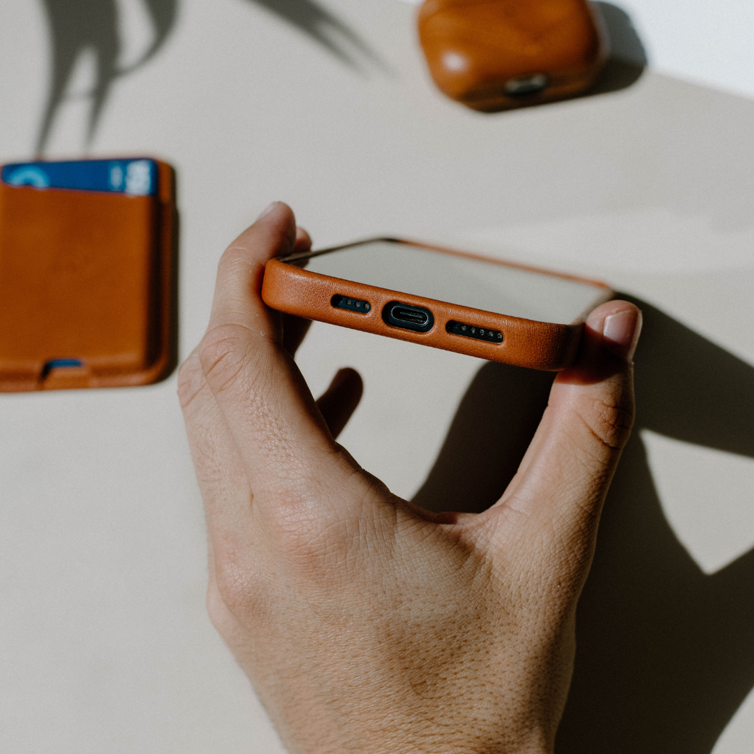 Hand holding sienna leather phone case showing bottom USB-C port and speaker cutouts