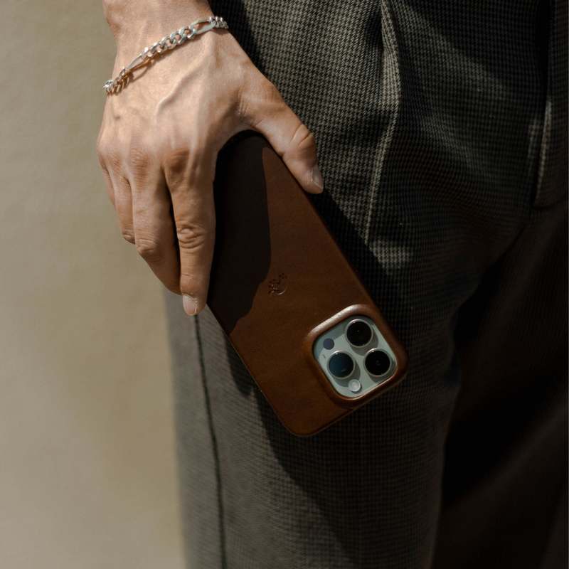 Hand wearing a silver chain bracelet holding a smartphone in a brown leather case against houndstooth trousers